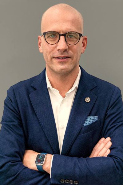 Foto di Carlo Apostoli a mezzo busto con braccia incrociate, camicia bianca, giacca blu su sfondo grigio